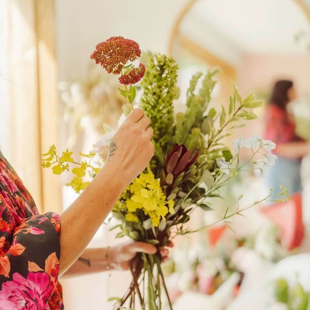 A hand places a flower stem inside of a bouquet while the other holds a bunch of colourful flowers together.